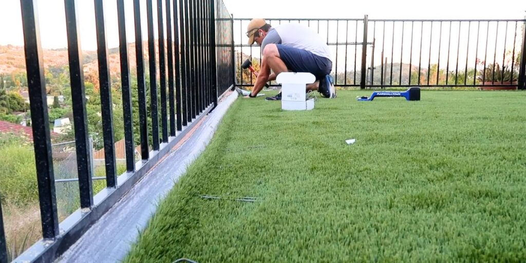 An installer kneels on a rooftop balcony, securing artificial grass near a black metal railing at sunset