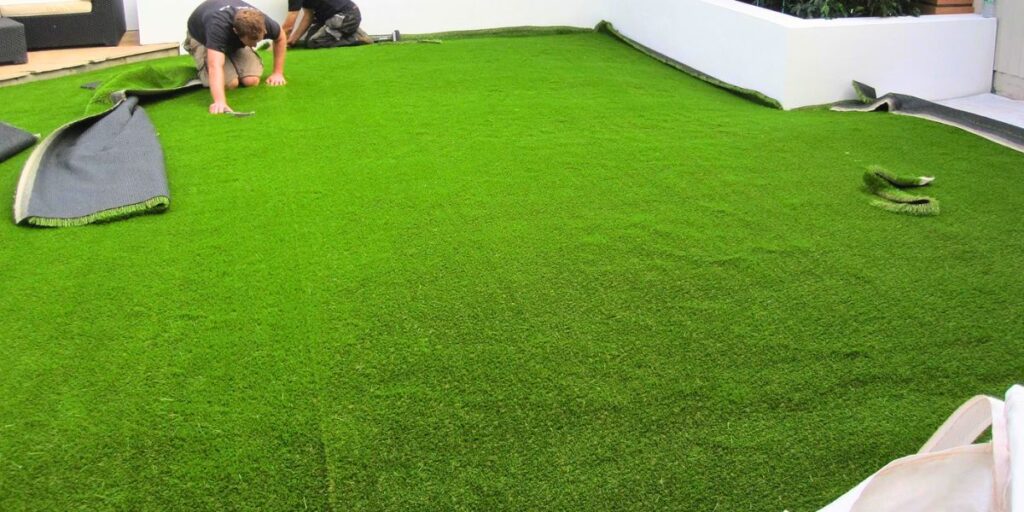 Two installers kneeling on a patio, rolling out and securing a large area of bright green artificial grass