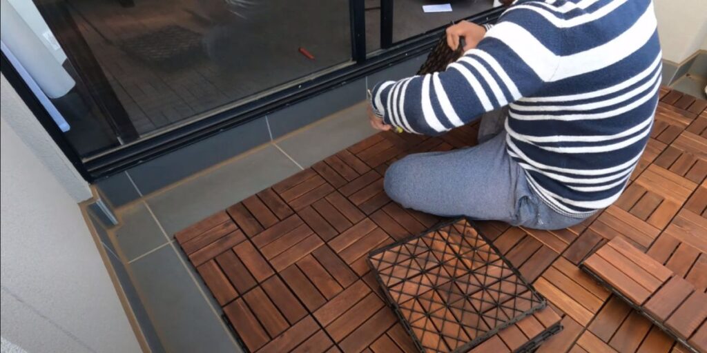 A person in a striped sweater installing dark brown interlocking wooden deck tiles on a balcony floor